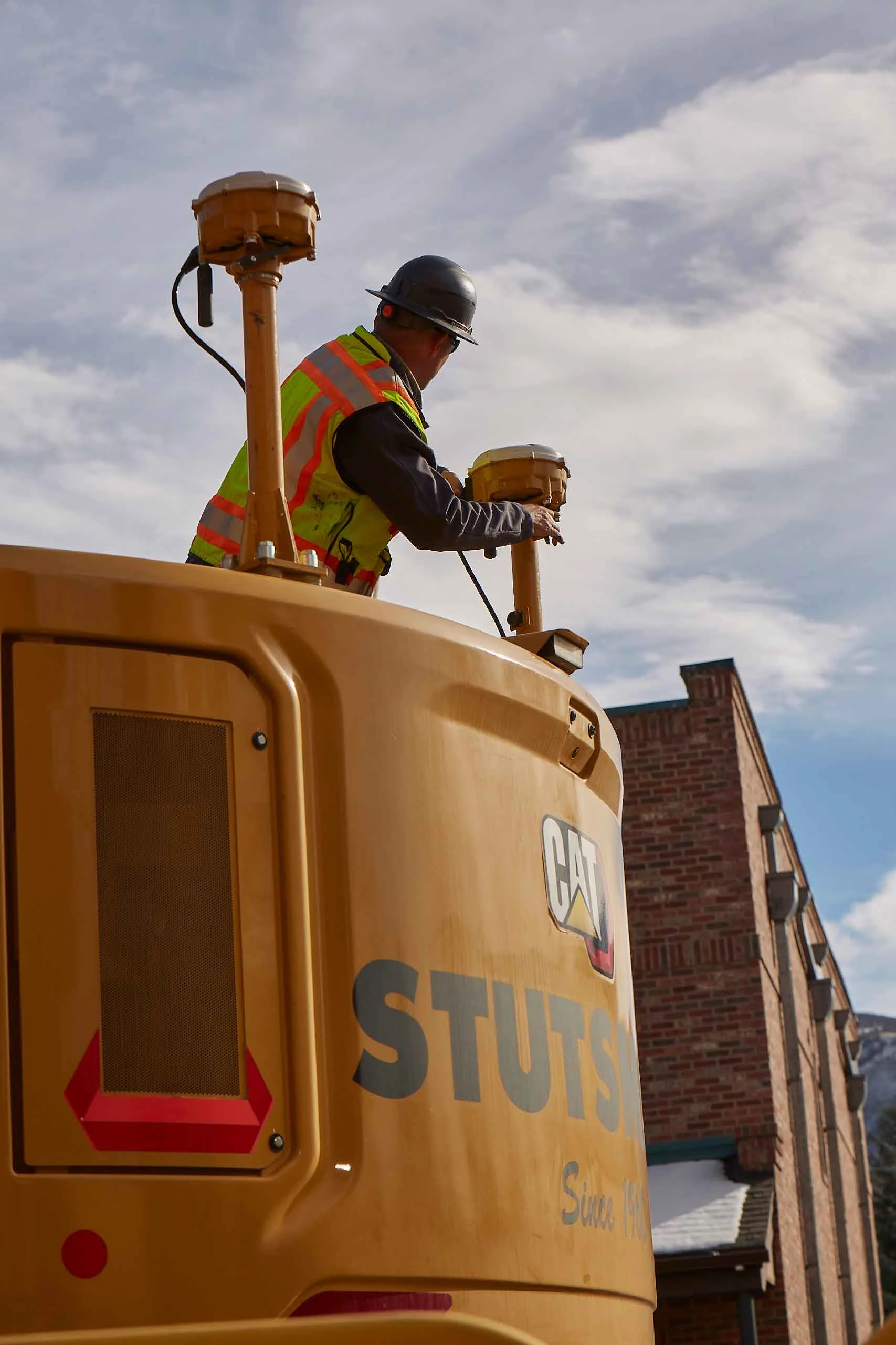A worker in a hard hat and safety vest adjusts a GPS receiver on a Caterpillar machine.