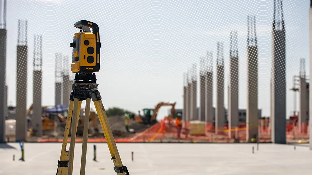 A yellow Trimble 3D laser scanner on a tripod at a construction site with concrete pillars and an excavator in the background.