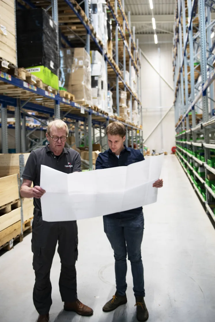 Two men in a warehouse examine a large blueprint together, surrounded by tall shelves of inventory.