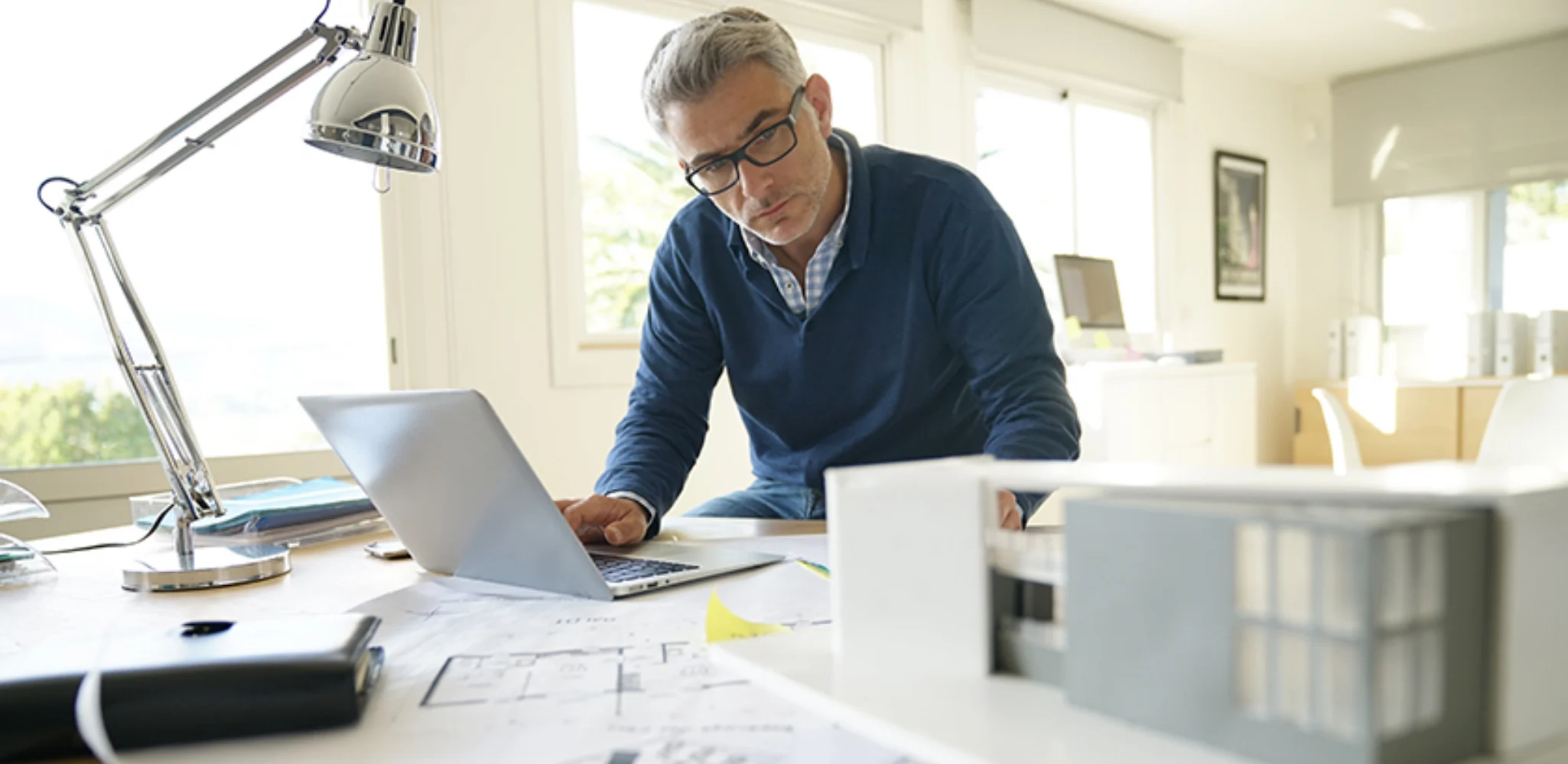 A middle-aged architect with glasses looks down at blueprints on his desk while using a laptop.