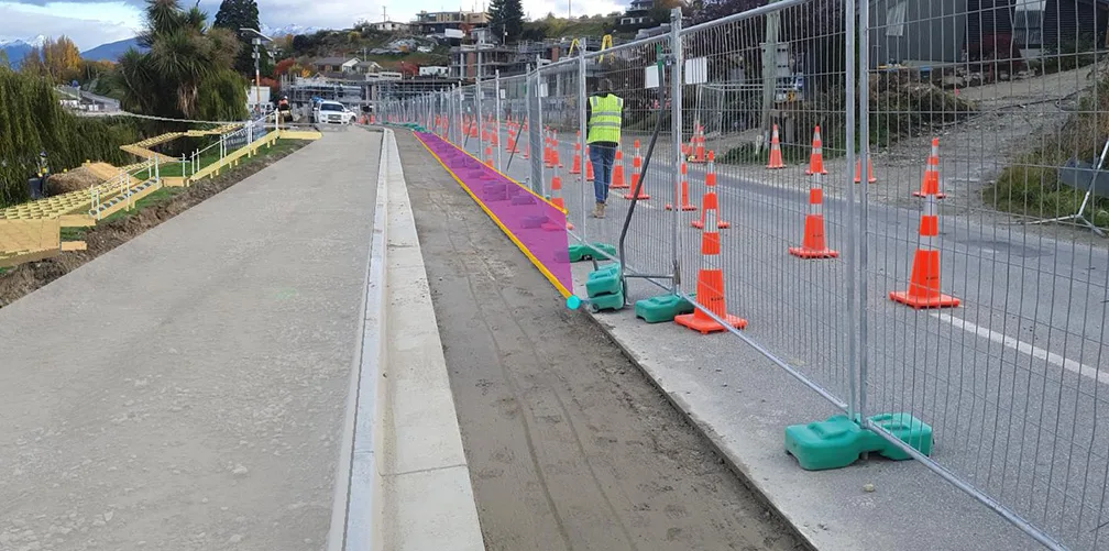 Construction site with safety fencing and orange traffic cones along a paved path.