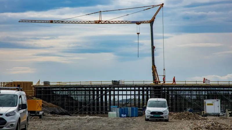 Construction site with a large crane and workers on a bridge under construction, with vans and equipment in the foreground.
