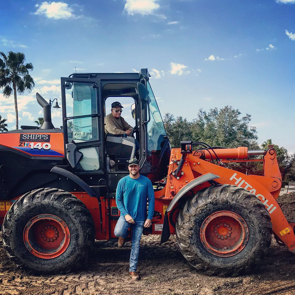 Two men pose with a Hitachi ZW140 loader. One man sits in the driver's seat, the other stands in front of the machine.