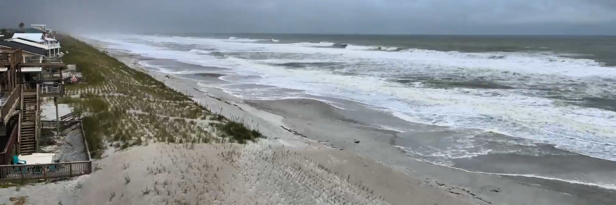 A wide shot of a beach before a storm, showing white-capped waves crashing onto the shore near coastal houses and sand dunes under a dark, heavy sky.