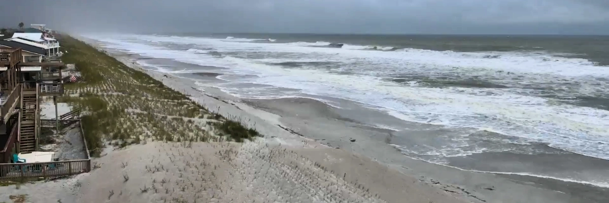 A wide shot of a beach before a storm, showing white-capped waves crashing onto the shore near coastal houses and sand dunes under a dark, heavy sky.