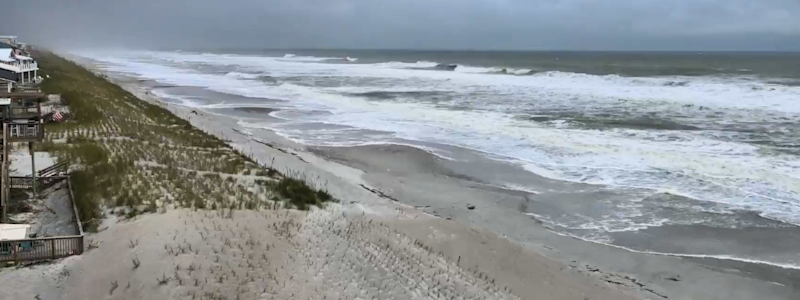 A wide shot of a beach before a storm, showing white-capped waves crashing onto the shore near coastal houses and sand dunes under a dark, heavy sky.