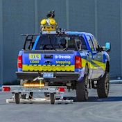 A blue and yellow TLS truck equipped with Trimble LiDAR mapping technology and sensors on the roof, towing a small trailer on a road.