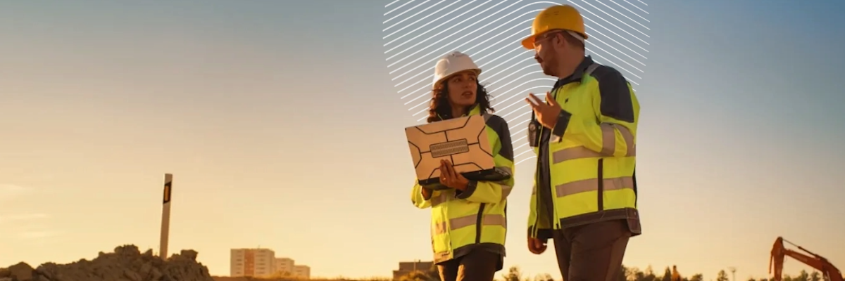 Workers in safety gear discussing plans at a construction site during sunset.