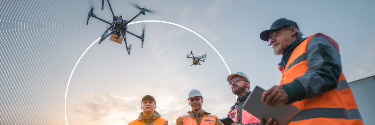 Four men in orange reflective vests in a circle watching one of them manage a drone in the sky above them.
