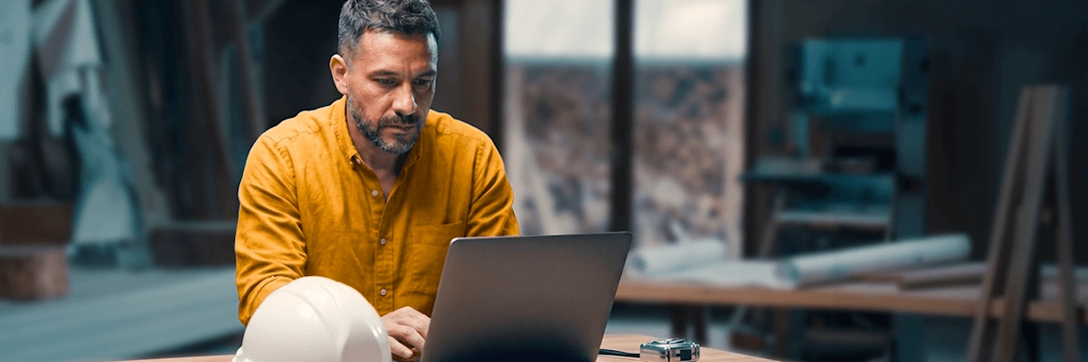 A focused contractor in a yellow shirt works on a laptop in a woodshop next to a white hard hat.