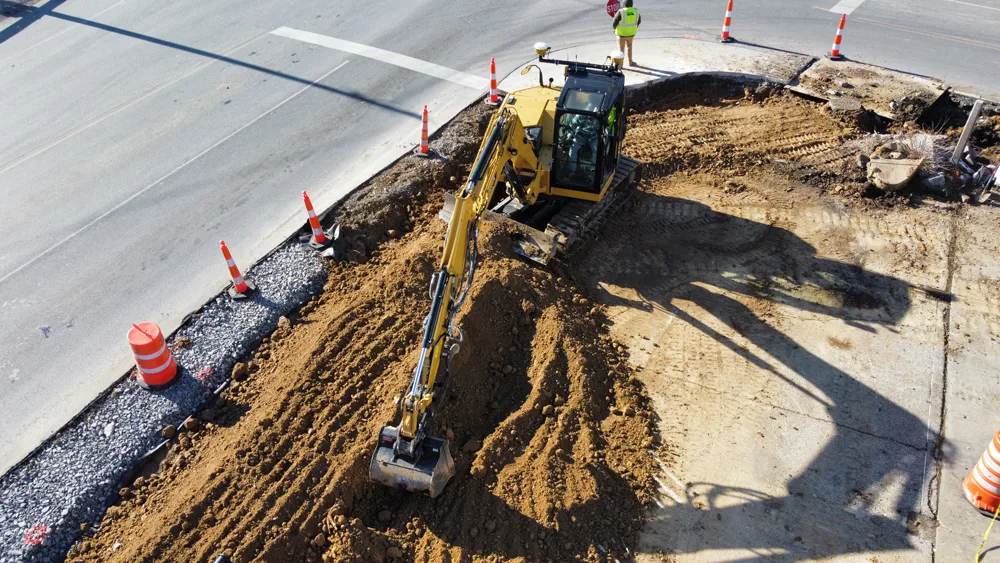 Yellow excavator digging a trench next to a road with orange traffic cones and gravel.