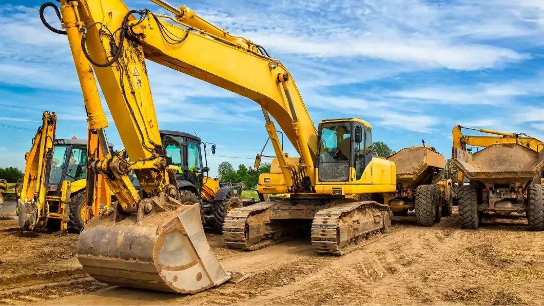 A large yellow excavator sits on a dirt construction site. Two dump trucks are parked behind it under a blue sky.
