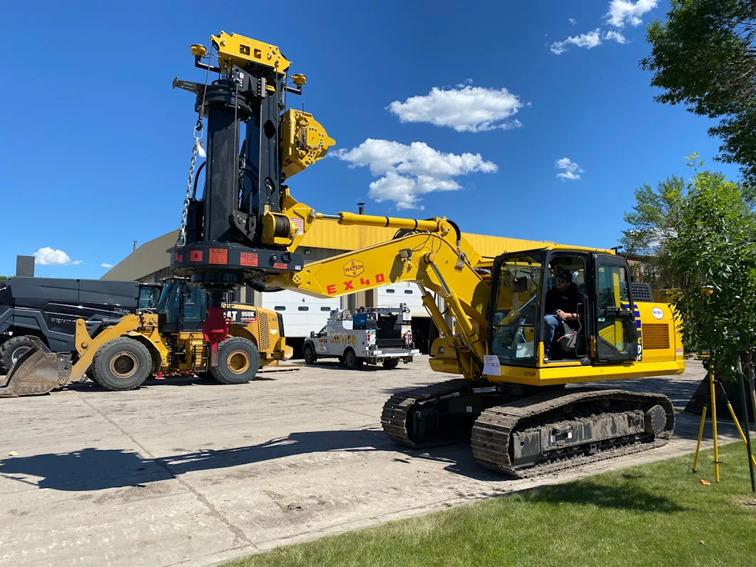 A yellow excavator is attached to a large black drilling attachment, with a wheel loader in the background.