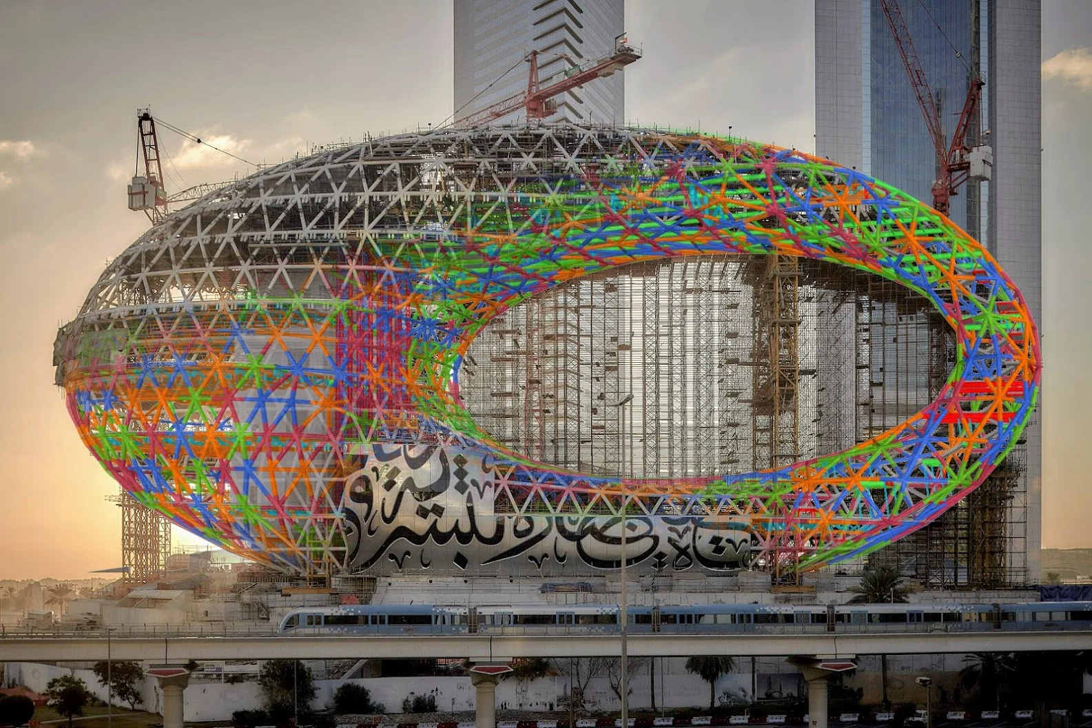 The Museum of the Future, under construction, designed with a steel exoskeleton covered in geometric patterns, set against modern skyscrapers.