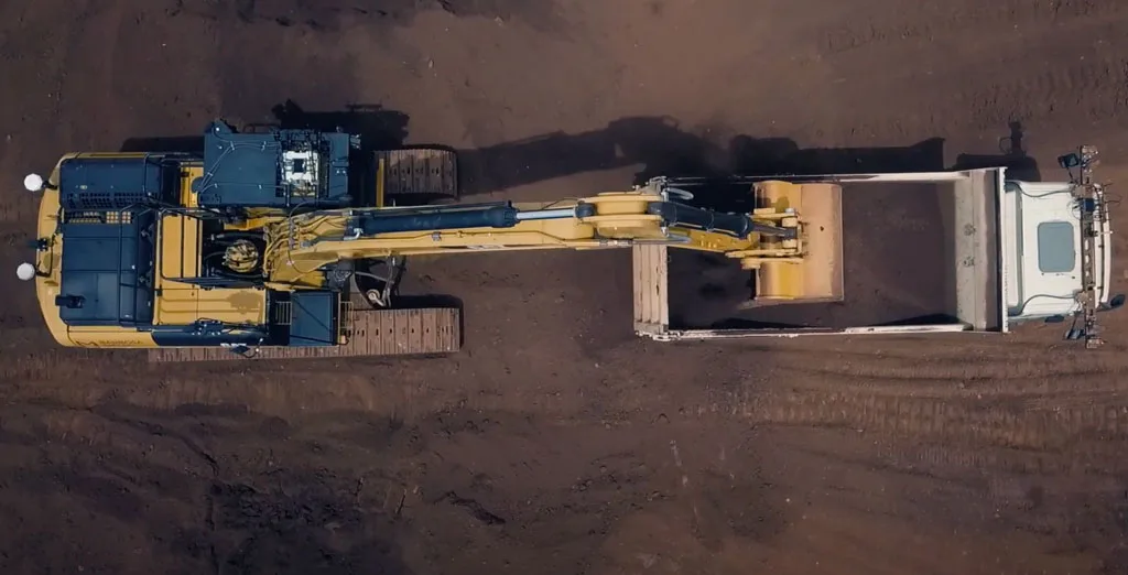 Yellow excavator loading dirt into a white dump truck from a bird's-eye view.