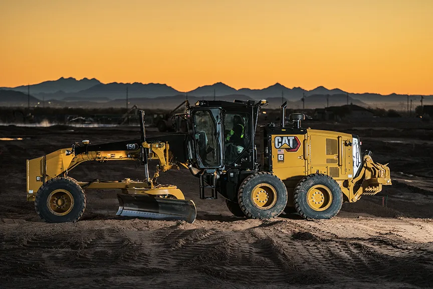 Yellow Caterpillar motor grader with operator, silhouetted against an orange sunset and distant mountains.
