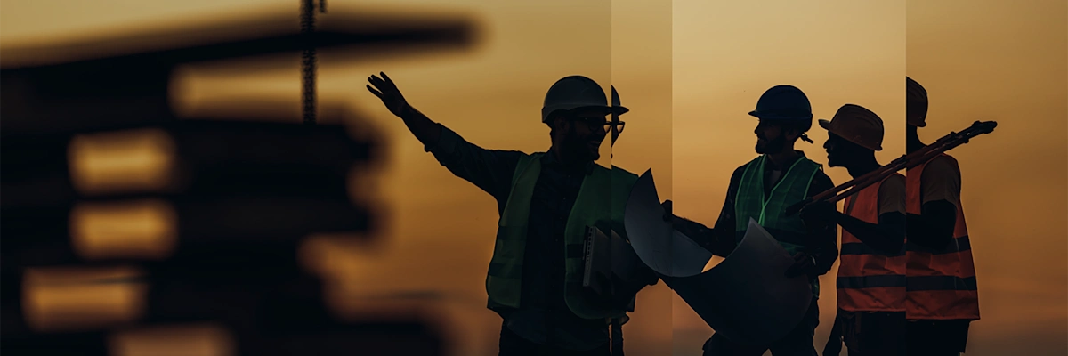 Three construction workers wearing hi-vis jackets and hardhats carry construction gear while conversating with a blurred constructing site in the background.