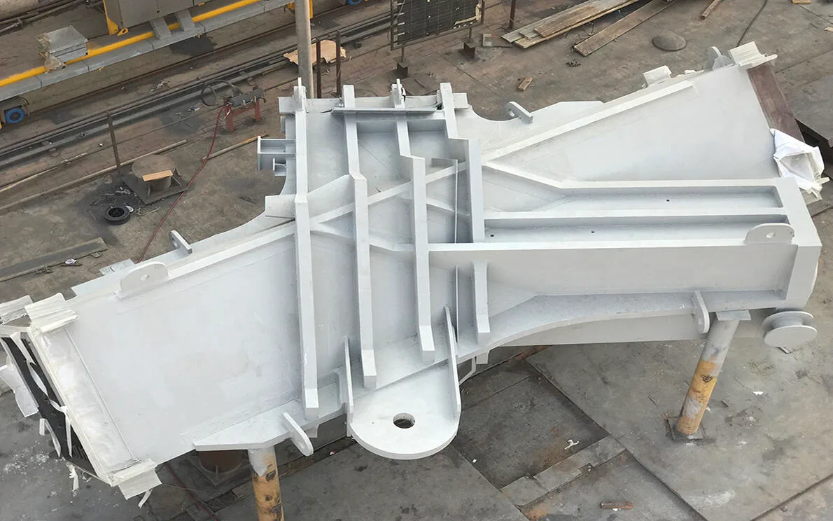 A large, gray, metal component with complex structural supports is shown from an elevated perspective in a workshop.
