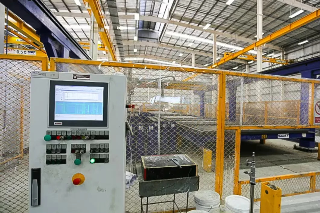 Factory control panel with a touchscreen display, set against a large industrial factory floor with overhead cranes and manufacturing equipment.