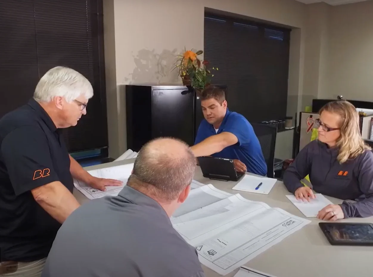 Four colleagues at a table review blueprints and digital plans.