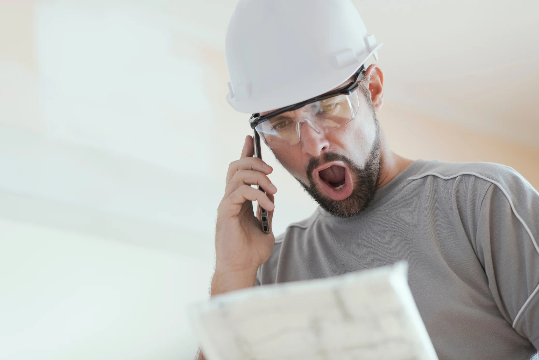 A construction worker in a hard hat and safety glasses looks shocked while talking on cell phone and holding a blueprint.