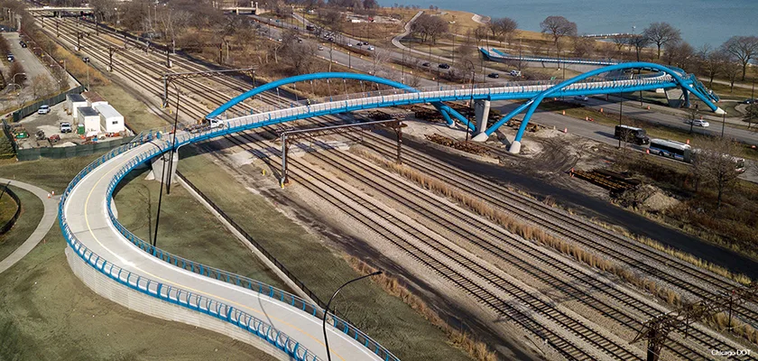 An aerial view of a pedestrian and bicycle bridge with a curved, wave-like design, spanning multiple train tracks with a body of water and a park.