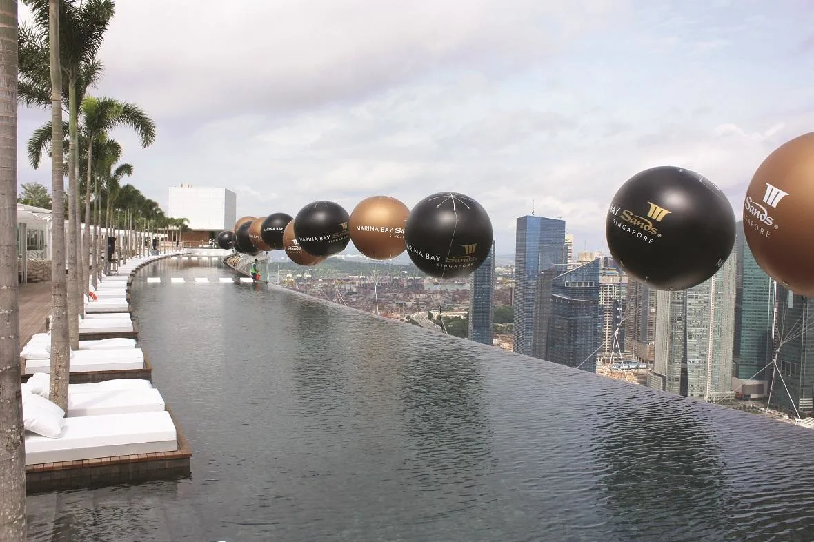 Large black and gold balloons float above the edge of an infinity pool overlooking a cityscape.
