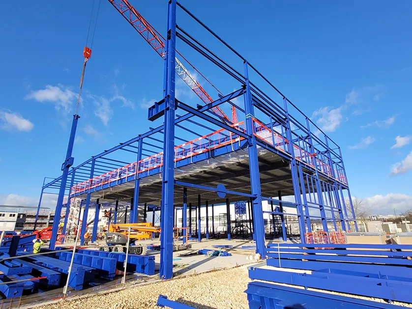 A large steel building frame, painted blue, is under construction on a sunny day with a crane overhead.