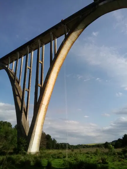 A lone person stands at the top of a massive concrete arch bridge, with a rope dangling below them into the green landscape.