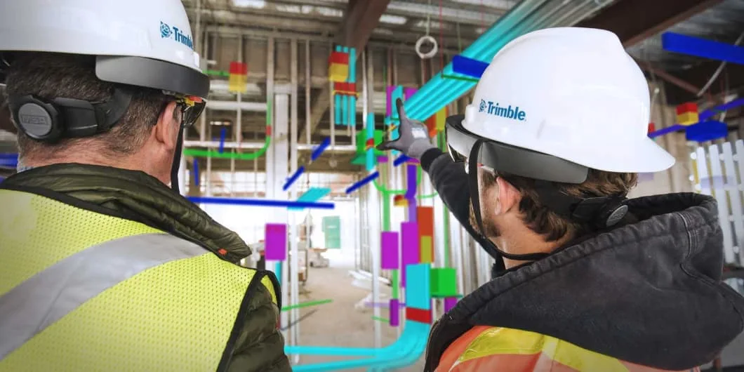 Two construction workers wearing hard hats and mixed reality headsets look at a digital model overlaid on a construction site.