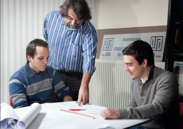 Three men huddle around a table covered with blueprints, with one man pointing at the plans.