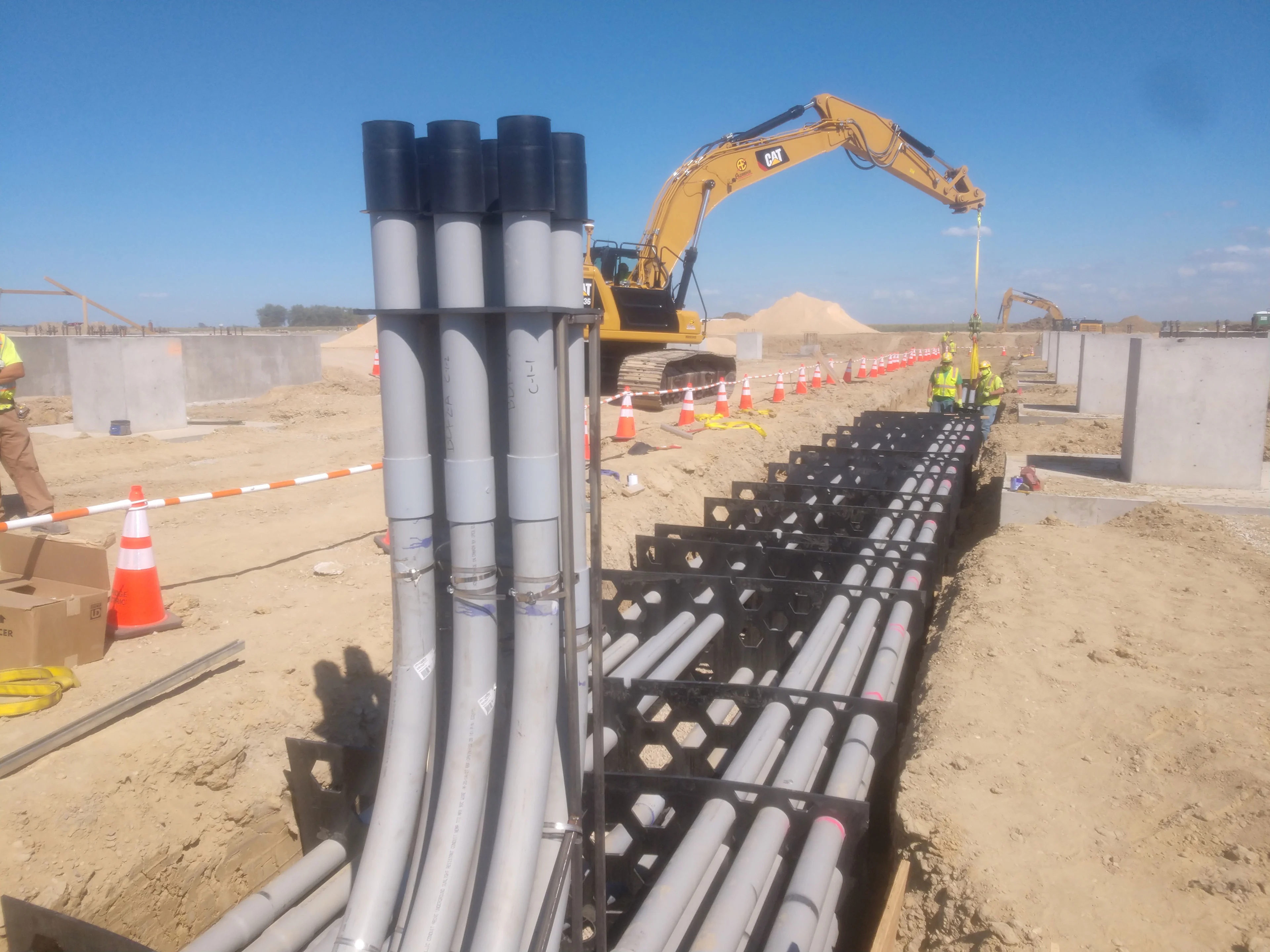 Construction site with workers installing conduit pipes with an excavator in the background.