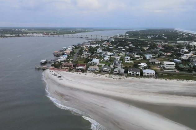 A wide shot of a beach before a storm, showing white-capped waves crashing onto the shore near coastal houses and sand dunes under a dark, heavy sky.