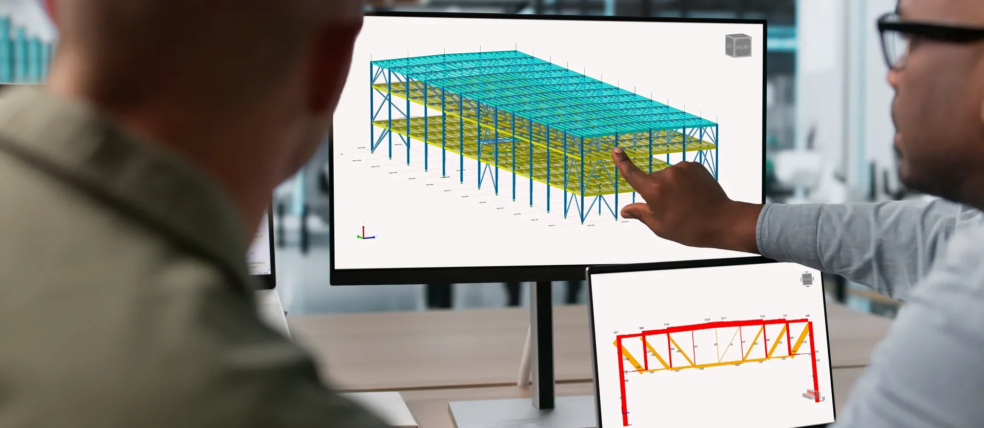 Over-the-shoulder view of two professionals collaborating in front of dual monitors; one person points to a turquoise and yellow 3D structural steel building model on the main screen while a secondary screen displays a red detailed cross-section.