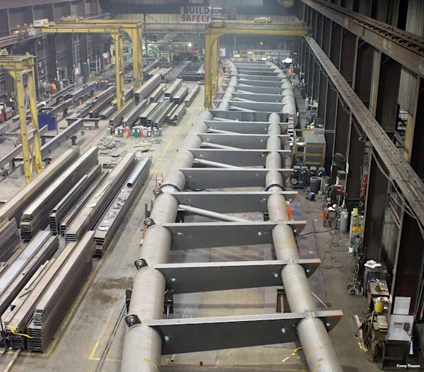 A long, segmented pedestrian bridge is under construction in a large industrial workshop, with steel beams and materials stacked nearby.