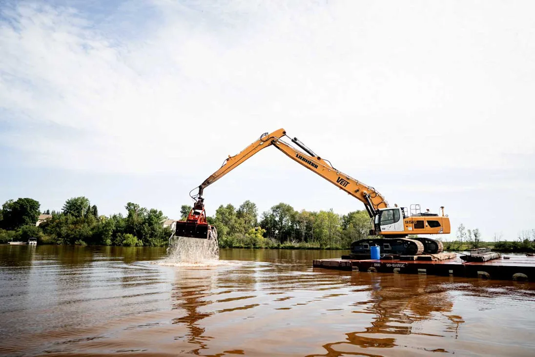 An excavator on a barge lifts a claw filled with water and sediment from a river.