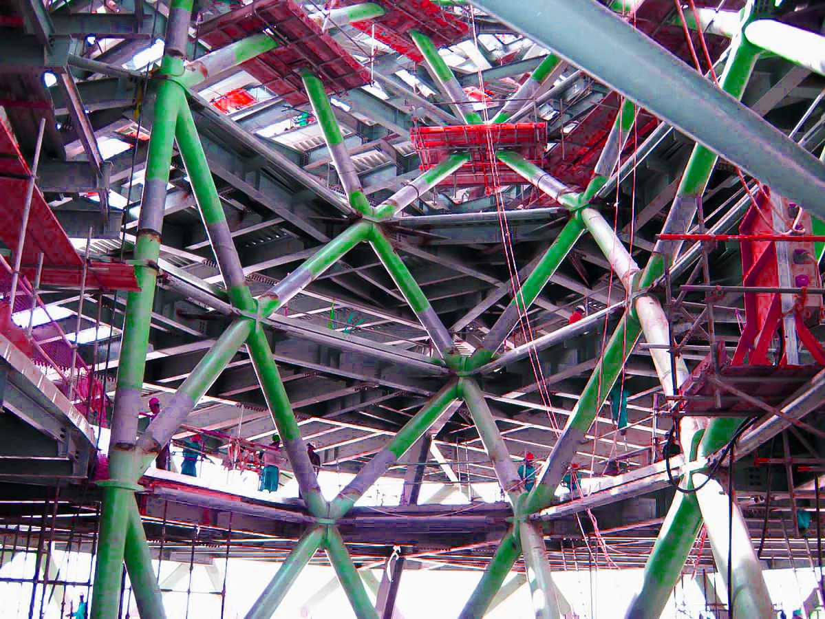 Close-up view of a complex steel structure under construction, with green and grey painted beams forming an intricate geometric pattern.