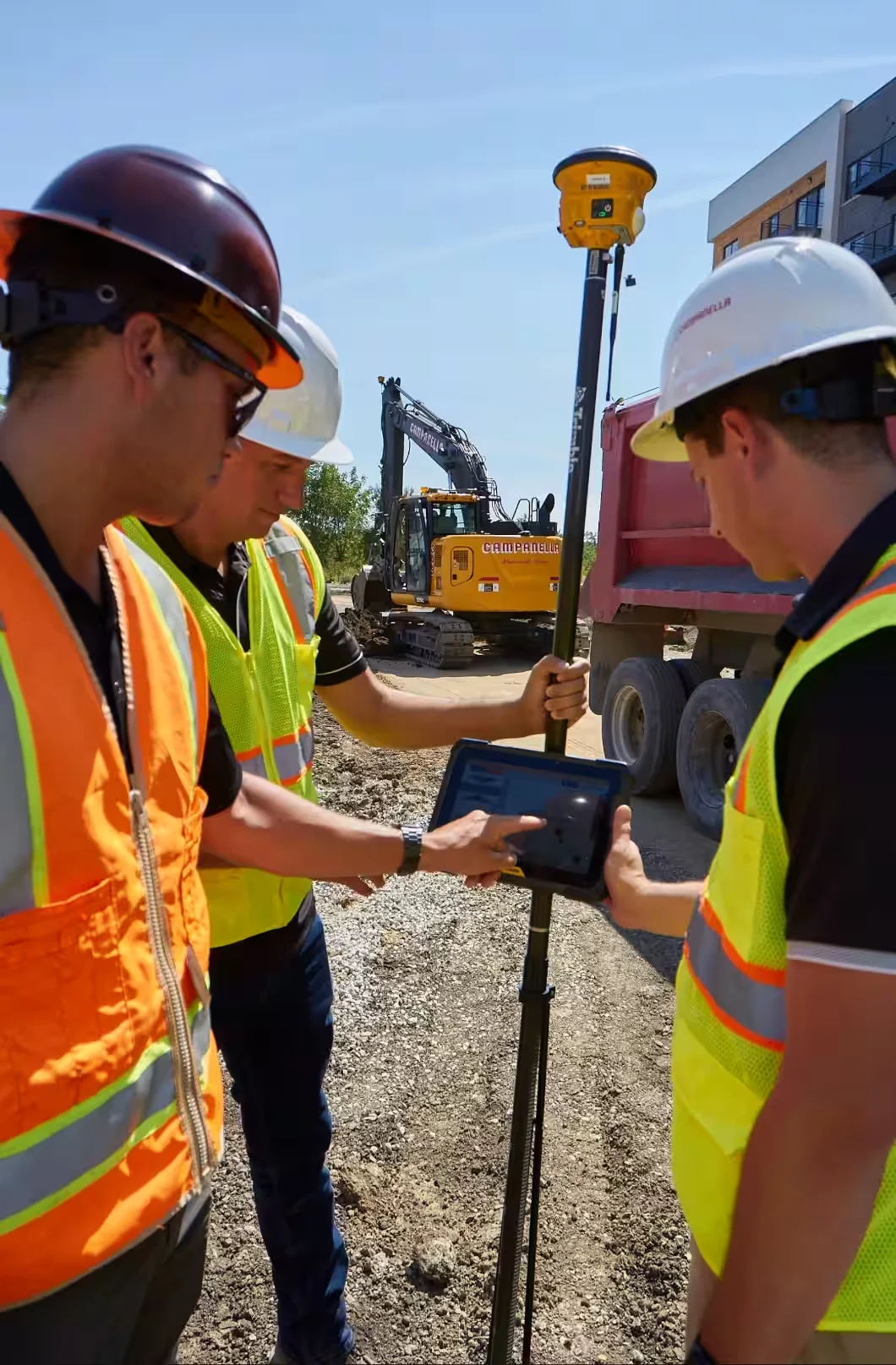 Three construction workers examine a tablet connected to a Trimble GPS surveying pole at a construction site.