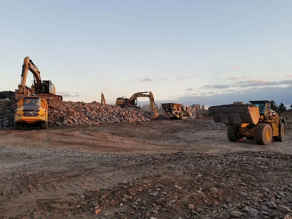 Construction vehicles including excavators and a loader operate at a rocky quarry site under a pale sky.