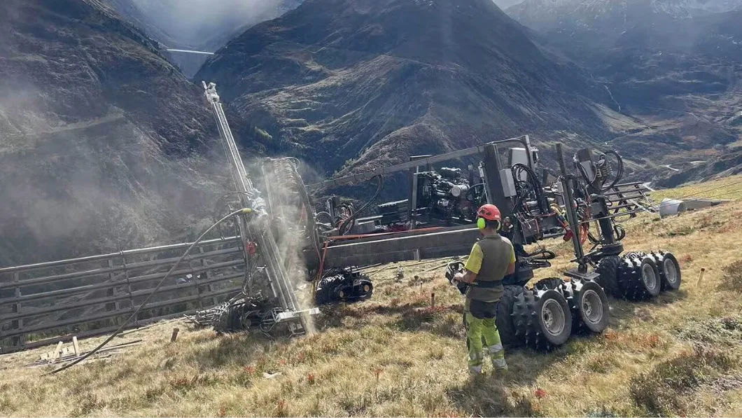Worker in safety gear operates heavy drilling machinery on a grassy mountain slope, with misty peaks in the background.