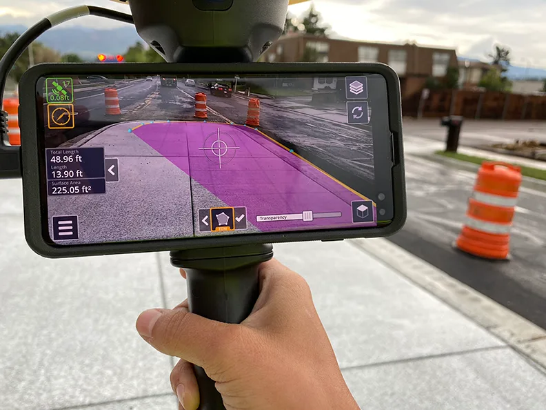 Smartphone displaying augmented reality overlay of construction site measurements with traffic cones in background.