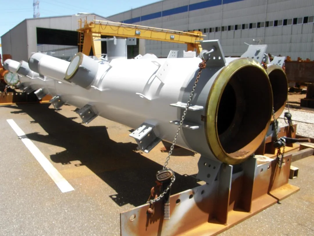 A large, white, cylindrical metal component rests on a yellow and brown industrial support structure outside a factory building.