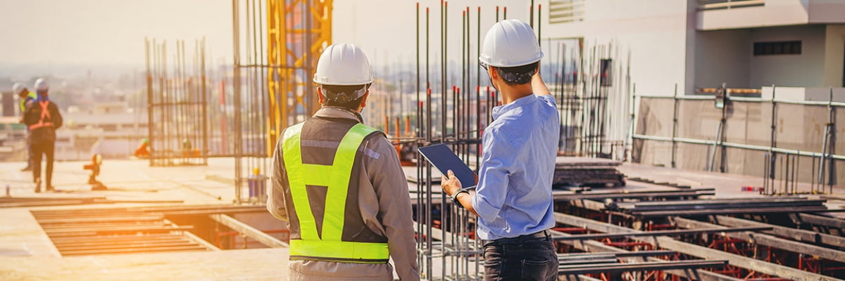 Two engineers in hard hats look over a construction site while one holds a digital tablet.