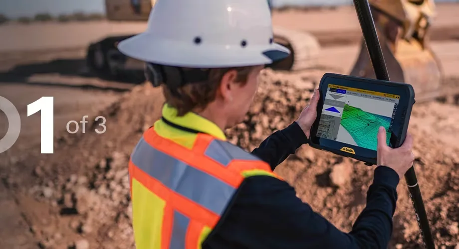 A construction worker in a hard hat and safety vest looks at a tablet displaying topographic data on a worksite.