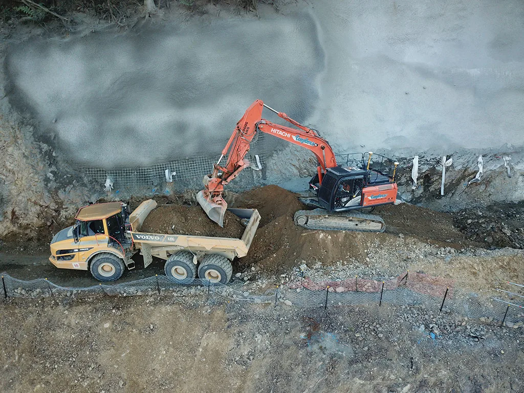 Construction vehicles, an excavator and a dump truck, work on a dirt excavation site.