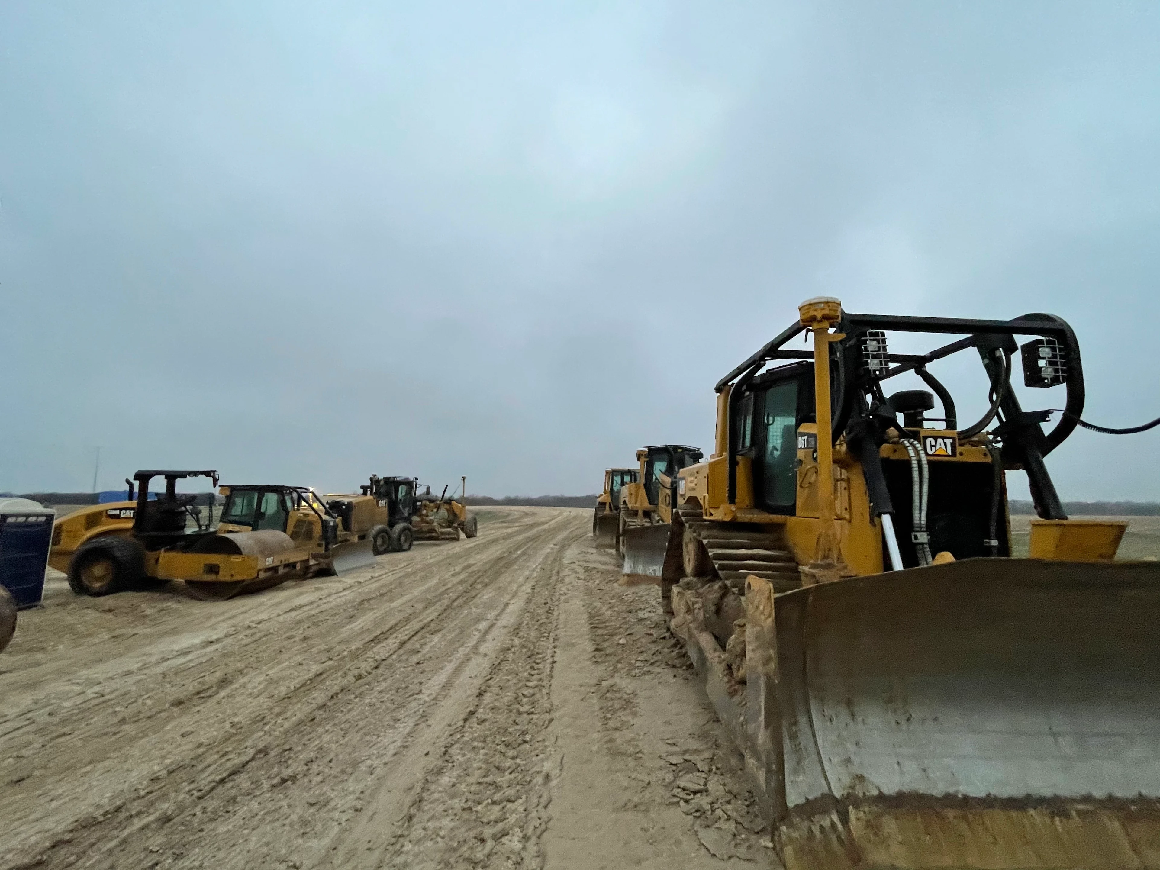 A fleet of yellow Caterpillar construction vehicles, including bulldozers and road rollers, are parked on a muddy construction site.