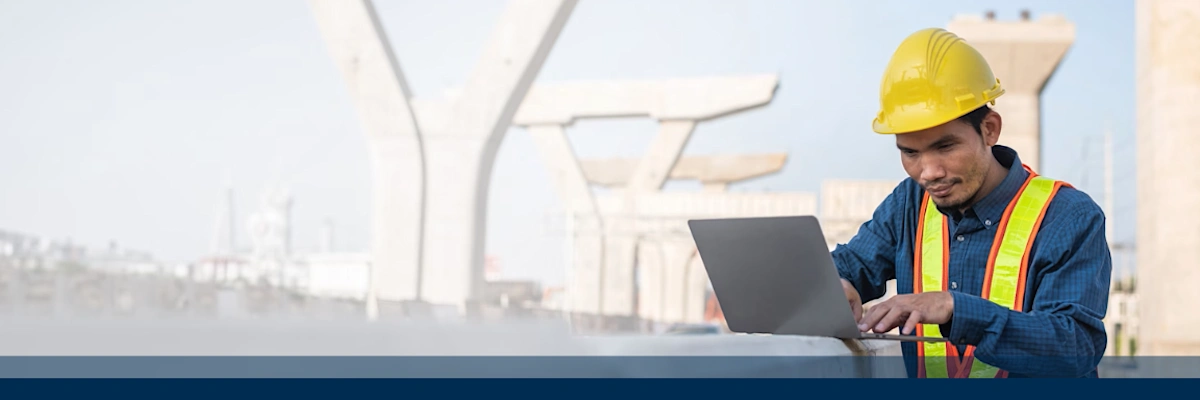 Construction worker in safety gear using a laptop at a bridge construction site.