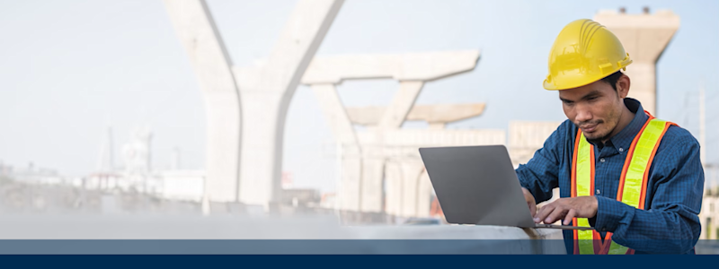 Construction worker in safety gear using a laptop at a bridge construction site.