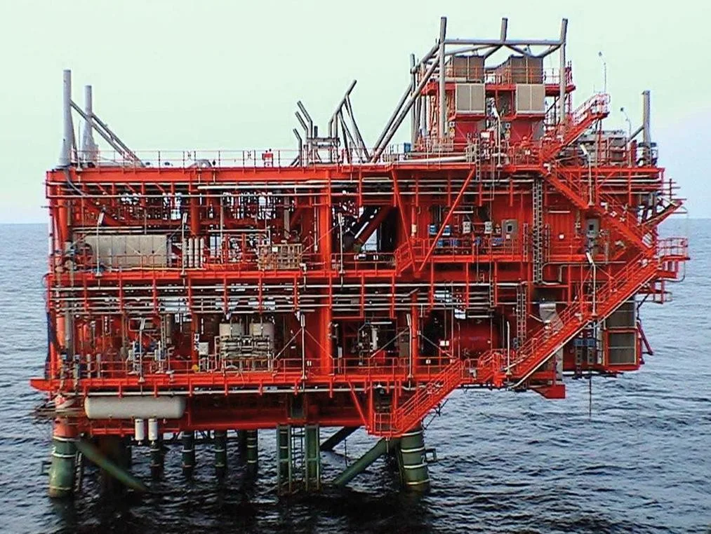 An offshore oil rig platform is painted bright red and stands on pilings in the ocean under a cloudy sky.