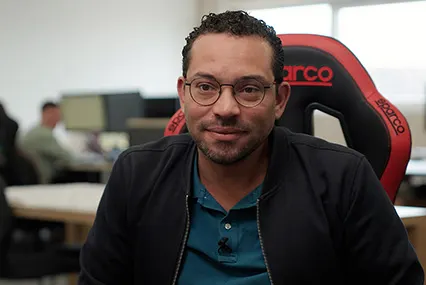 Person with glasses in black jacket and blue shirt sitting in a red gaming chair in an office environment.